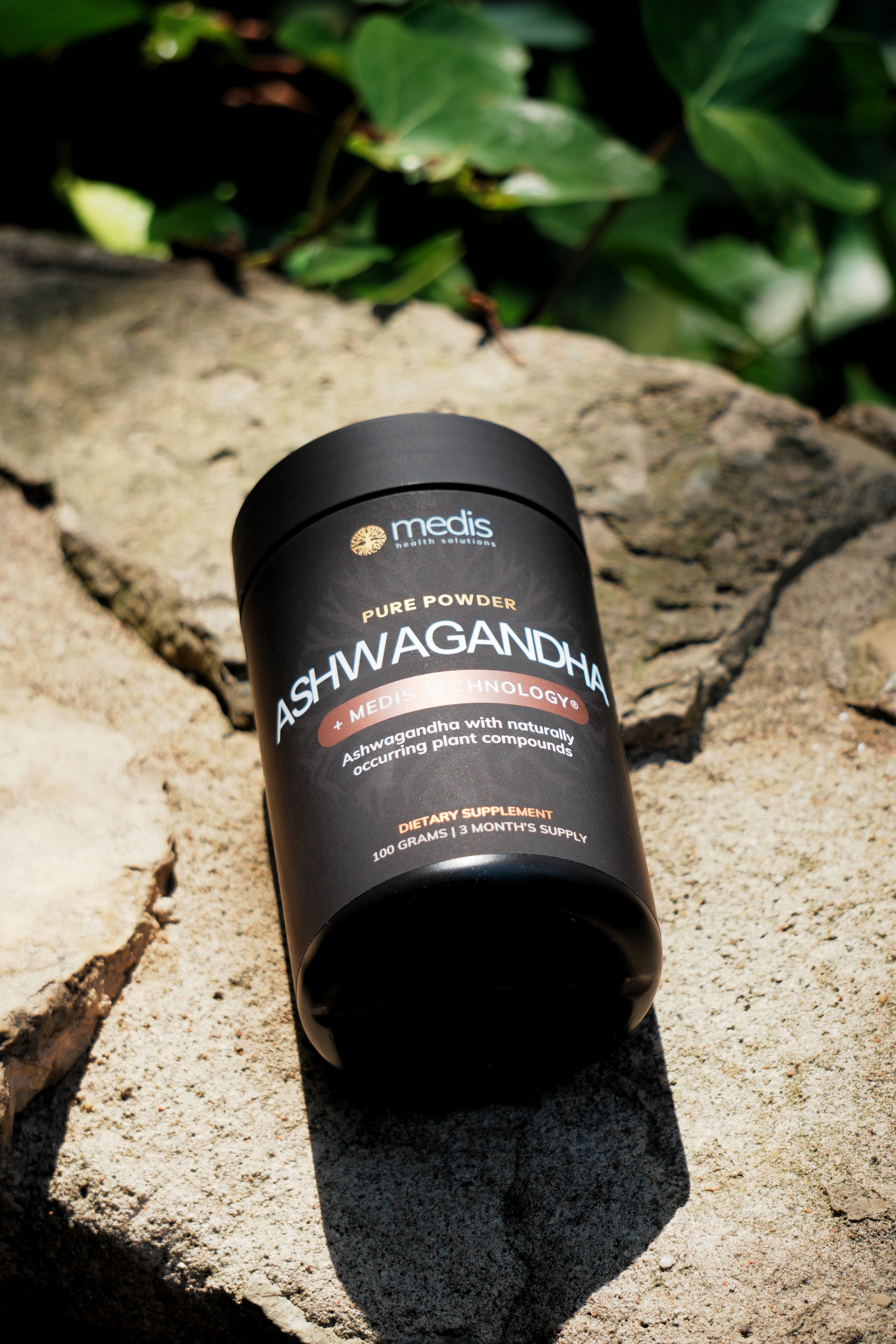 A black cylindrical tub of Medis Ashwagandha Pure Powder dietary supplement, placed on a textured stone surface with green foliage in the background.