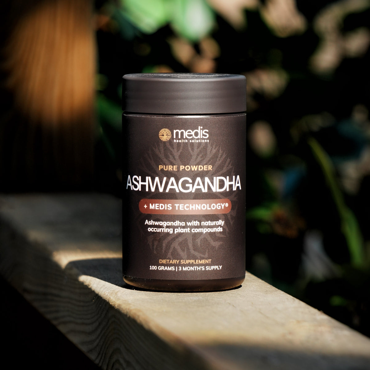 A dark brown jar of Medis Ashwagandha pure powder sits on a wooden ledge with a blurred natural background.