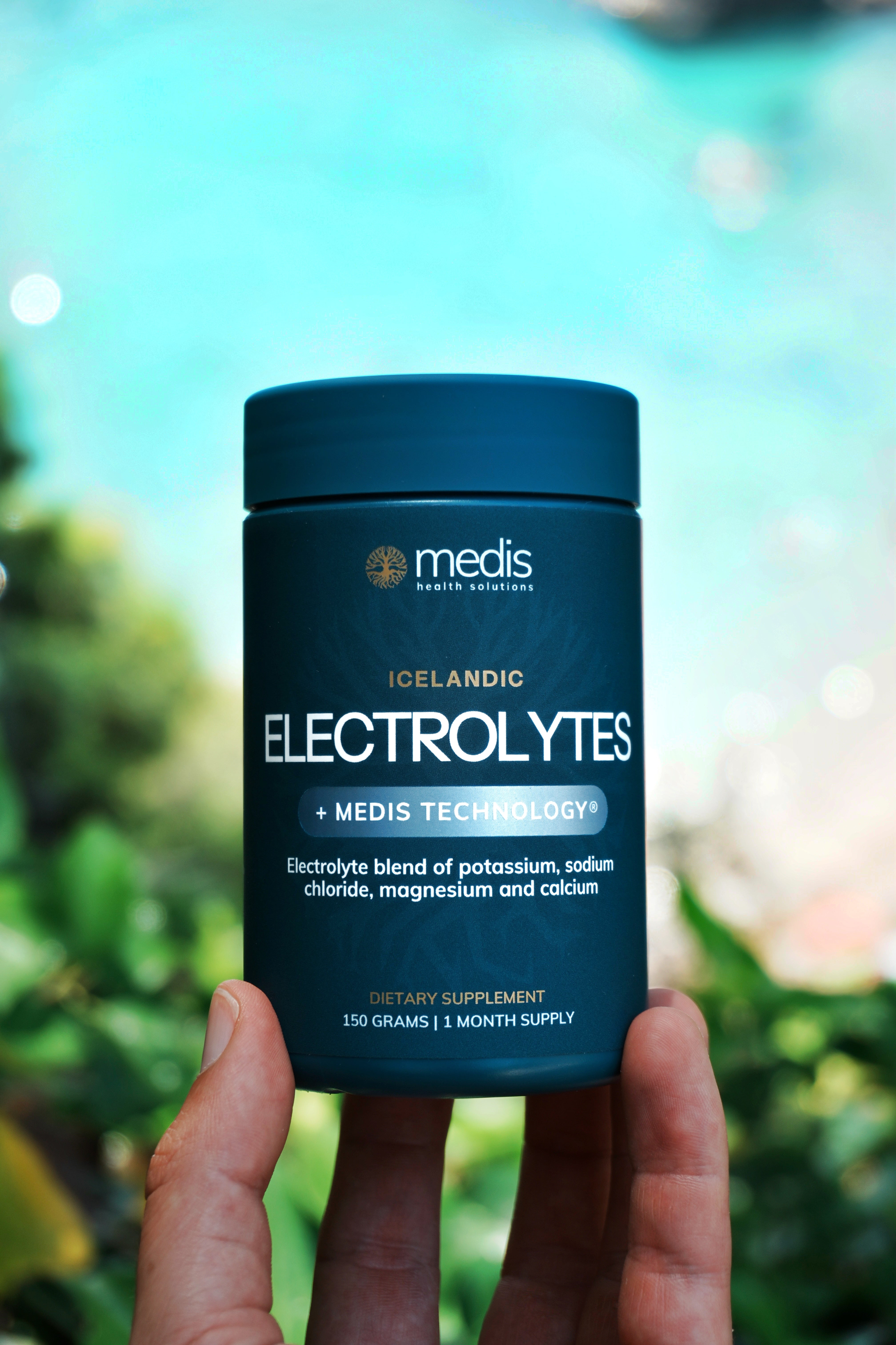 A hand holding a dark teal bottle of Medis Electrolytes + Medis Technology dietary supplement, with a blurred outdoor background of green foliage and bright blue water.
