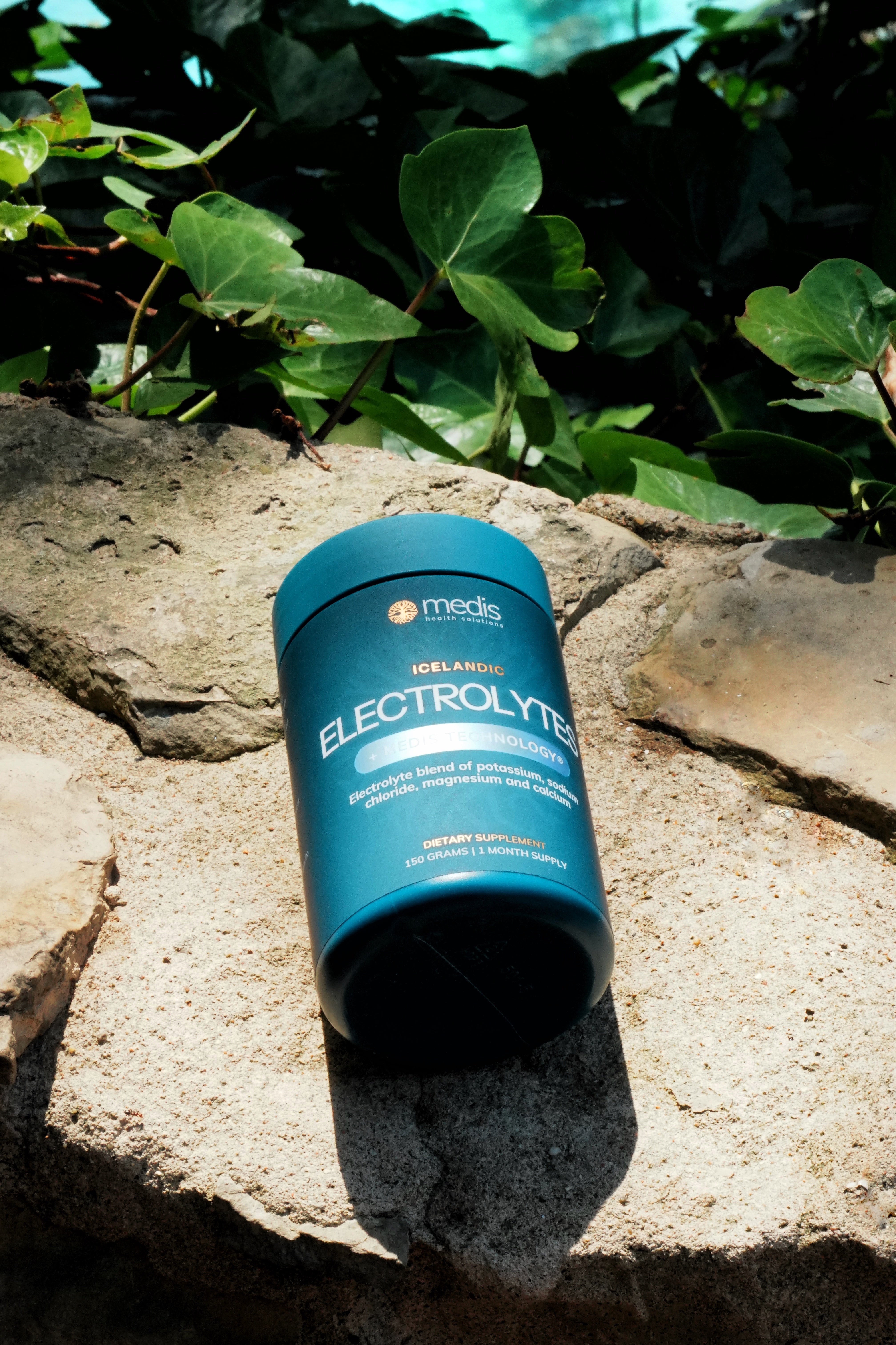 A blue cylindrical bottle of Medis Ionic Energy Electrolyte supplement placed on a stone surface with green ivy leaves in the background.