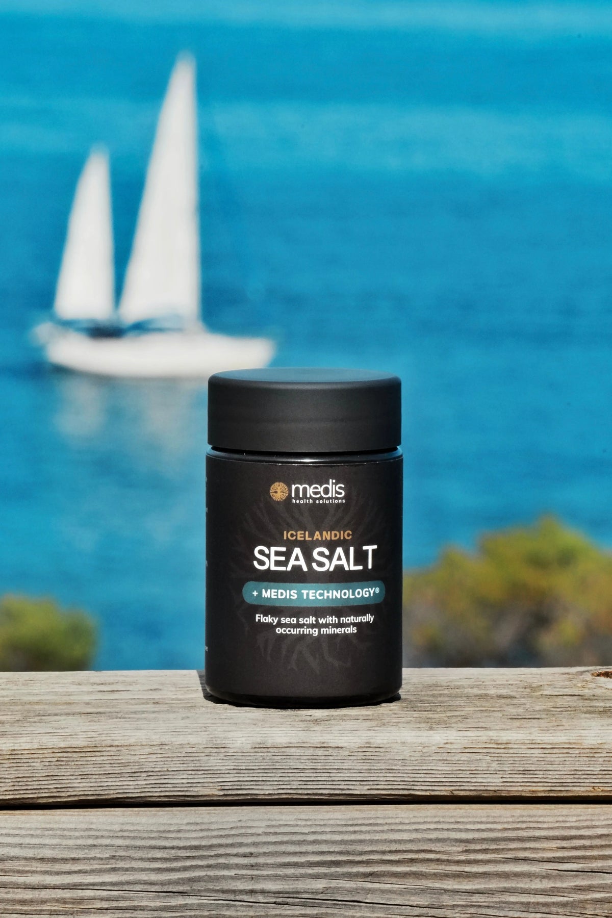 A black jar of Medis Iceland Salt sits on a weathered wooden deck with a blurred blue ocean and sailboat in the background.